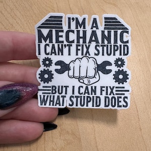 May include: A white sticker with black text that reads "I'M A MECHANIC I CAN'T FIX STUPID BUT I CAN FIX WHAT STUPID DOES." The design includes a wrench held by a fist and gear icons.