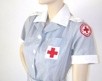 May include: A gray and white uniform dress with a white collar and short sleeves. The dress has a red cross on the chest and a red circle with a red cross and the text "American Red Cross Volunteer" on the right shoulder.