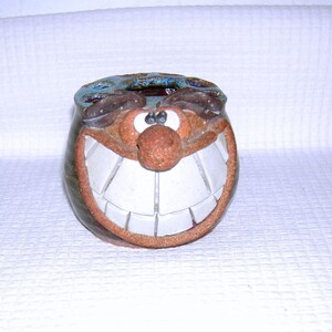 May include: A whimsical ceramic container with a smiling face design. The container features a brown face with a large white-toothed grin, a red nose, and blue accents around the top. The container is approximately 4 inches tall.