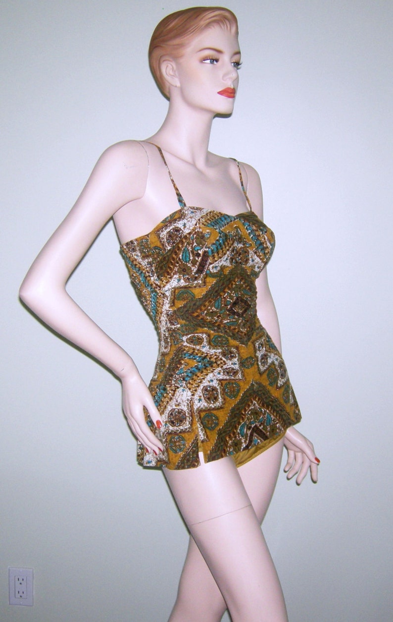 May include: A vintage-style swimsuit with a brown, gold, and blue geometric pattern. The swimsuit has a halter top and a skirt-like bottom.
