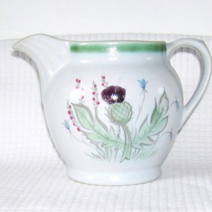 May include: A white ceramic creamer with a green rim and a painted design of a thistle flower with green leaves and purple and red accents.