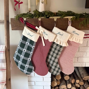 Puede incluir: Cuatro calcetines de Navidad personalizados colgados en fila. El primero es de cuadros verdes con pequeños árboles y el nombre Dad. El segundo es de cuadros rojos con borde festoneado y el nombre Mom. El tercero es de cuadros verdes con volante y el nombre Jack. El cuarto es de rayas rojas y blancas con volante y el nombre Remi.