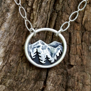 May include: A silver necklace with a circular pendant featuring a detailed silhouette of a mountain range with trees. The pendant is attached to a silver chain.