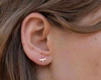 May include: A close-up of a person's ear with a small gold bird earring. The earring is a simple design with a delicate outline.