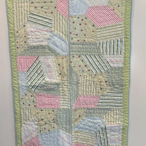 May include: A patchwork baby quilt with a green border and blue trim. The quilt is made of various fabrics in shades of pink, blue, and green with stripes and floral patterns.
