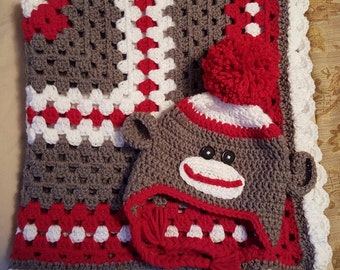 Puede incluir: Un juego de manta y gorro de bebé hechos a mano con ganchillo. La manta presenta un diseño de cuadrados de la abuela en gris, blanco y rojo. El gorro está diseñado para parecerse a un mono calcetín, con ojos de botón y un pompón rojo.
