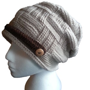 Può includere: Un berretto di lana beige con una fascia in crochet marrone e un bottone marrone. Il berretto è morbido e ha un motivo a treccia.