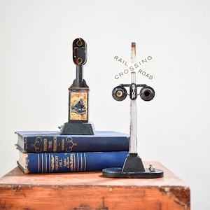 May include: A vintage toy train crossing signal with a black and white sign that reads "RAIL CROSSING ROAD" and a red "STOP" sign. The signal is on a wooden base and is next to a toy train signal with a black and white label that reads "The Heritage".