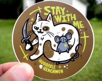 May include: A brown and white cat with a knife and a small gray mouse with a heart on its chest. The cat is holding the mouse and the text "Stay with me" is written in yellow on a brown background. The text "Huggable Henchmen" is written in yellow at the bottom of the sticker.