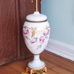 May include: A white ceramic table lamp with a gold-colored metal base and accents. The lamp features a floral design in pink and gold. The lamp has three light sockets and a cord with a plug.