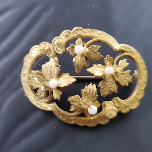 May include: A gold-toned brooch with a design of four leaves with a pearl in the center of each leaf.