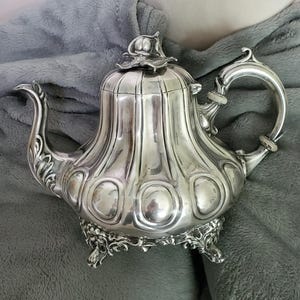 May include: A silver-colored teapot with a decorative floral design on the lid and base. The teapot has a curved spout and handle, and a ribbed body. The teapot is sitting on a gray blanket.