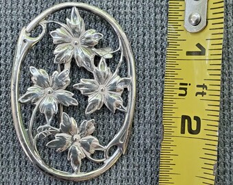 May include: A silver oval brooch with a floral design. The brooch features four stylized flowers with intricate details and a delicate vine pattern.