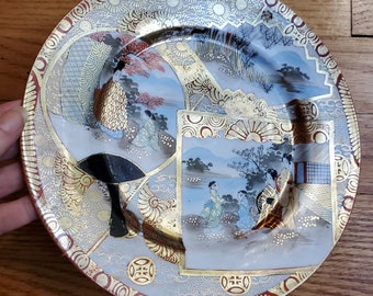 May include: A white porcelain plate with a gold and red rim. The plate features a Japanese-inspired design with figures in traditional clothing and a landscape scene. The plate is decorated with gold leaf and intricate details.