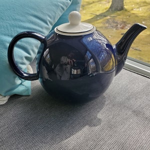 May include: A dark blue teapot with a white lid and a white rim around the top. The teapot has a round body and a curved spout.