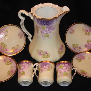 May include: A vintage tea set with a cream-colored pitcher, four cups, and four saucers. The set features a floral design with purple and pink flowers and gold accents.