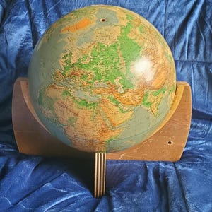 May include: A vintage globe with a detailed map of the world, mounted on a wooden stand. The globe features a classic color palette with shades of green, brown, and blue. The stand has a curved design and a vertical support.