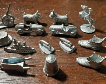 May include: A collection of 12 vintage silver-toned game pieces, including a thimble, a boot, a wheelbarrow, a ship, an iron, a car, a dog, a hat, a cannon, and a cowboy on a horse.