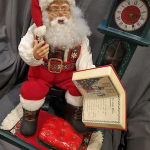 May include: A Santa Claus figurine wearing a red suit, hat, and boots, with a white beard and glasses. He is holding a book with the text "The Night Before Christmas". A green clock is in the background.