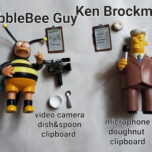 May include: A yellow and black bee figurine with a camera, a white dish, a spoon, and a clipboard. A brown figurine of Ken Brockman with a microphone, a donut, and a clipboard.