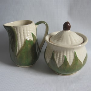 May include: A ceramic creamer and a lidded sugar bowl set featuring a textured corn cob design. Both pieces are cream-colored with green painted husks at the base. The sugar bowl has a matching lid with a small brown knob on top.