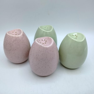 May include: Four ceramic salt and pepper shakers in an egg shape. Two are light pink with dark speckles, and two are light green. Each shaker has a series of small holes at the top for dispensing.