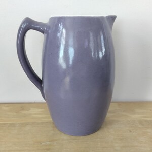 May include: A lavender-colored ceramic pitcher with a curved handle and a spout. The pitcher has a smooth, glossy finish and a white interior rim. It is sitting on a wooden surface.