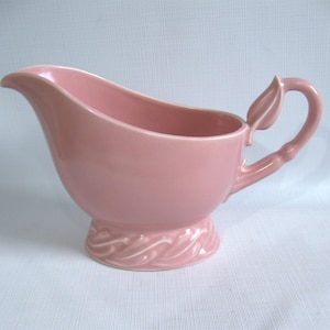 May include: A vintage pink ceramic gravy boat. The boat features a curved spout, a handle with a leaf detail, and a decorative base. The gravy boat is a soft pink color and is likely from the mid-20th century. It is a classic piece of kitchenware.