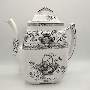 May include: A white ceramic teapot featuring black transferware patterns. The design includes a central urn, a basket of flowers, and geometric borders. It has a square-shaped body, a curved handle, a spout, and a lid with a decorative finial.