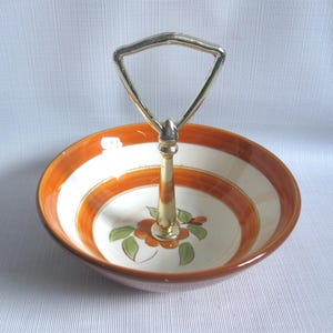May include: A ceramic serving bowl with a gold-colored handle. The bowl features a white interior with orange and brown accents and a floral design. The handle is triangular in shape.