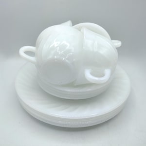 May include: A stack of white ceramic teacups and saucers. The cups have curved handles and sit atop matching saucers with a spiral design. The set is all white, creating a clean and classic aesthetic. The teacups and saucers are stacked on top of each other.