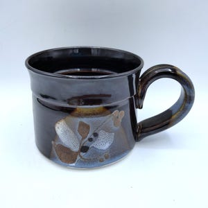 May include: A ceramic mug with a dark brown exterior and a matching handle. The mug features a decorative floral design in shades of brown, white, and blue. The mug is approximately 4 inches tall and 3.5 inches in diameter.