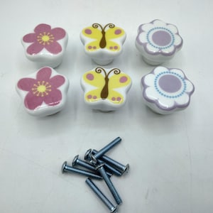 May include: Six ceramic cabinet knobs arranged in two rows, featuring two pink flower designs, two yellow butterfly designs, and two purple flower designs. Below the knobs are six silver-colored metal screws with flat heads for installation.