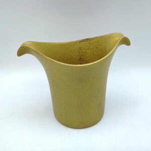May include: A yellow-green ceramic vase with a flared, wavy rim. The vase has a cylindrical body and a textured surface. The rim curves outward, creating a unique, organic shape. The vase is set against a white background.