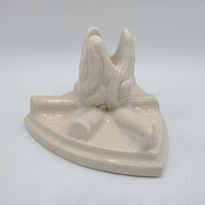 May include: Off-white ceramic iron rest with a stylized design. The rest features a raised, curved handle area and a flat, iron-shaped base. The design includes a decorative bow and a textured surface.