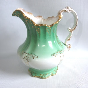 May include: A vintage ceramic pitcher with a light green and white gradient. The pitcher features gold trim along the rim, base, and handle, with ornate gold detailing on the body. The handle is white with gold accents.