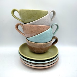 May include: A stack of six speckled ceramic cups and saucers in various colors. The cups are stacked on top of each other, with the saucers at the bottom. Colors include green, gray, pink, blue, and brown. The cups have curved handles.