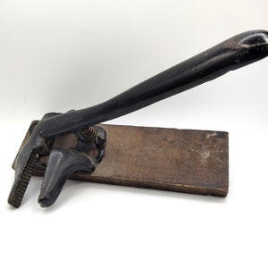May include: An antique black metal tool with a long handle and a hinged clamp, mounted on a rectangular wooden base. The tool appears to be a vintage press or clamp, likely used for a specific craft or trade.