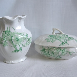 May include: A white ceramic vase and lidded dish set. Both pieces feature a green floral design. The vase has a ruffled rim, and the dish has a handle on the lid. The set is likely vintage or antique.