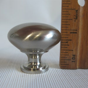 May include: A brushed nickel cabinet knob with an oval top and a decorative base. The knob is approximately 1.5 inches tall. The knob is next to a wooden ruler.