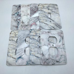May include: A collection of marble-patterned light switch and outlet covers. The covers feature a white base with gray and pink veining, offering a natural stone aesthetic. Designs include single switch and double outlet configurations.