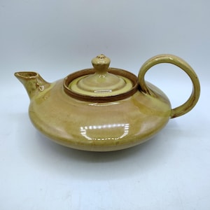 May include: A low-profile, wide-bodied ceramic teapot with a glossy, honey-yellow glaze. It features a short, pointed spout, a curved handle, and a matching circular lid with a small, rounded knob handle centered on top of the vessel.