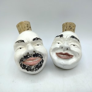 May include: Two white ceramic bottle stoppers with human face designs. One has a black beard and mustache, the other has a smiling expression. Both have cork tops.