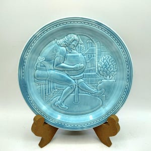 May include: A light blue ceramic plate with a raised relief design of a potter at work. The plate features a detailed scene with a potter, buildings, and a tree. The plate is displayed on a wooden stand.