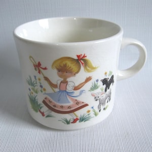 May include: White ceramic mug featuring a whimsical illustration of a girl in a blue dress with a red bow, holding a wand. The design includes a black lamb, a white lamb, and colorful flowers. The mug has a curved handle.