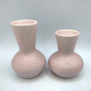 May include: Two light pink ceramic salt and pepper shakers with a speckled finish. The taller shaker has a slender neck and a bulbous base, while the shorter shaker features a wider neck and a rounded body, both with holes on top.
