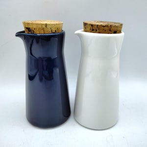 May include: Two ceramic cruet bottles, one navy blue and one white, each topped with a cork stopper. The bottles have a slender, curved design with a pouring spout. The background is a plain white.