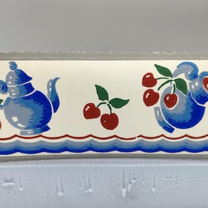 May include: A rectangular, white container with a gray rim and two metal handles. The container is decorated with blue teapots and red cherries. A blue and red wave pattern runs along the bottom.