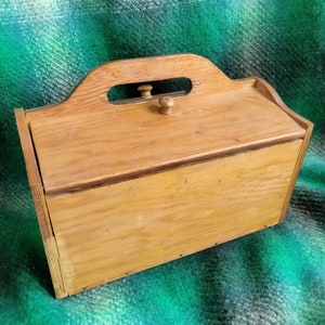 May include: A light brown wooden box with a handle and a small knob on the lid. The box has a rectangular shape with a slightly angled top. The wood grain is visible, and the box appears to be vintage.