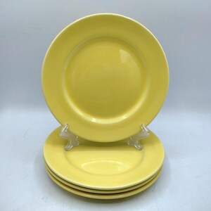 May include: A stack of four yellow ceramic plates. One plate is propped up on a clear plastic stand, with three additional plates stacked below. The plates have a simple, round design and a glossy finish.